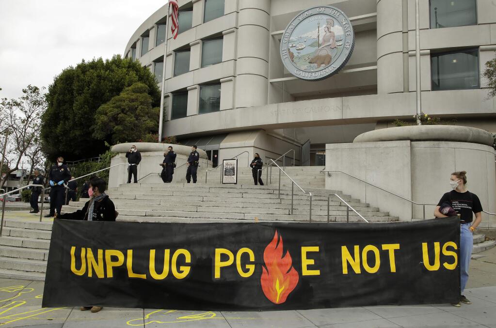 Masked protesters hold a banner in front of law enforcement officers at the State of California Public Utilities Commission building on Wednesday, May 20, 2020, in San Francisco. Activists drew yellow chalk outlines of PG & E fire victims (seen lower left) outside the building and demanded California Governor Newsom cancel a plan that would bring the failed utility out of bankruptcy. (AP Photo/Ben Margot)