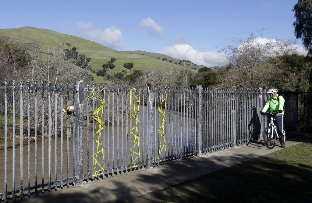 A bicyclist sits near flowers placed on a fence as he looks toward Alameda Creek near Fremont, Calif., Tuesday, Jan. 24, 2017. An unidentified woman's car plunged into rushing waters of Alameda Creek after colliding with another vehicle on Niles Canyon Road on Saturday, Jan. 21. (AP Photo/Jeff Chiu)