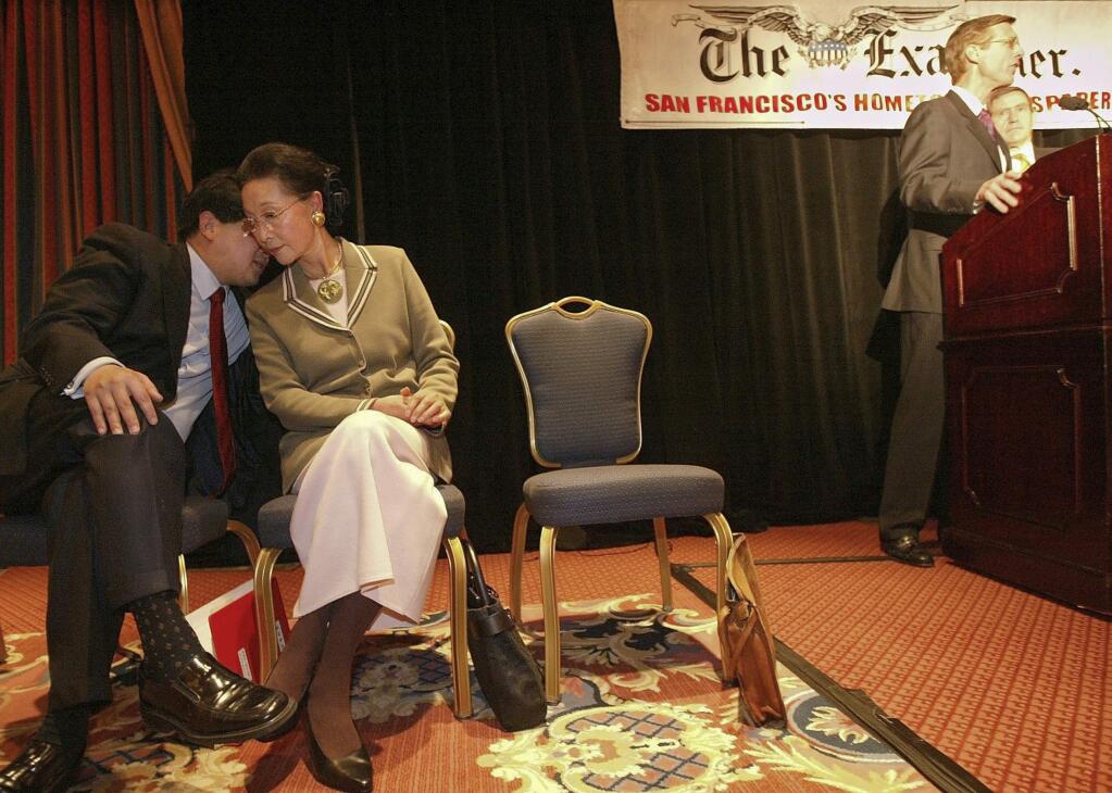 FILE - In this Feb. 19, 2004, file photo, James Fang, left, talks with his mother, Florence Fang, as president and publisher of The San Francisco Examiner and Independent newspapers Scott McKibben, second from right, announces The Anschutz Co. purchasing the newspapers from the Fang family at the Ritz-Carlton in San Francisco. A San Francisco Bay Area town is suing Florence Fang, the owner of the quirky Flintstone House, alleging she violated local codes when she put dinosaur sculptures in the backyard and made other landscaping changes that caused local officials to declare it a public nuisance. The town of Hillsborough filed a complaint this week against Fang, the media mogul who purchased the orange-and-purple, bulbous-shaped house in 2017. (AP Photo/Jeff Chiu, File)