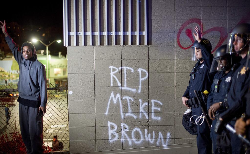 A protester stands near a wall with a graffiti and police officers in Oakland, Calif., on Monday, Nov. 24, 2014, after the announcement that a grand jury decided not to indict Ferguson police officer Darren Wilson in the fatal shooting of Michael Brown, an unarmed 18-year-old. Several thousand protesters marched through Oakland with some shutting down freeways, looting, burning garbage and smashing windows. (AP Photo/Noah Berger)