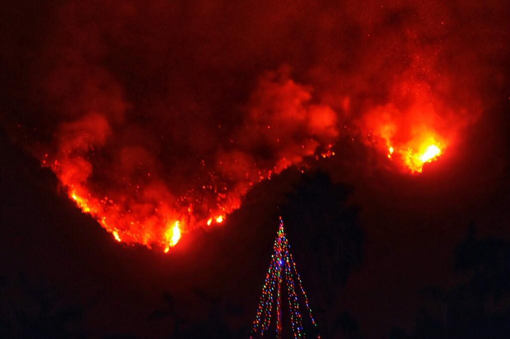 In this photo provided by the Santa Barbara County Fire Department, with flames burning behind it, a Christmas tree stands as a lone sentinel in the front yard of an evacuated home in Carpinteria, Calif., Monday, Dec. 11, 2017. (Mike Eliason/Santa Barbara County Fire Department via AP)