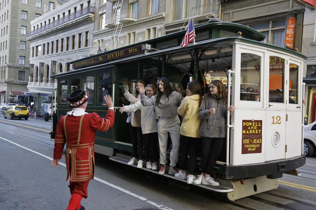 In this photo taken Monday, Jan. 6, 2020, a group of girls riding a Powell Street cable car are greeted by Beefeater doorman Tom Sweeney outside the Sir Francis Drake Hotel in San Francisco. Another bit of old, quirky San Francisco will be no more when Tom Sweeney finally hangs up his white pressed collar and signature Beefeater jacket after more than four decades as chief doorman at downtown's Sir Francis Drake Hotel. Sweeney has opened doors for movie stars and shaken hands with every U.S. president since Gerald Ford, with the exception of Donald Trump. He's taken photos with countless visitors from around the world, often after telling them where to catch the cable car and how to get to Fisherman's Wharf. The man known as a 'San Francisco original' and 'living landmark' retires Sunday, Jan. 12, 2020, after 43 years. (AP Photo/Eric Risberg)