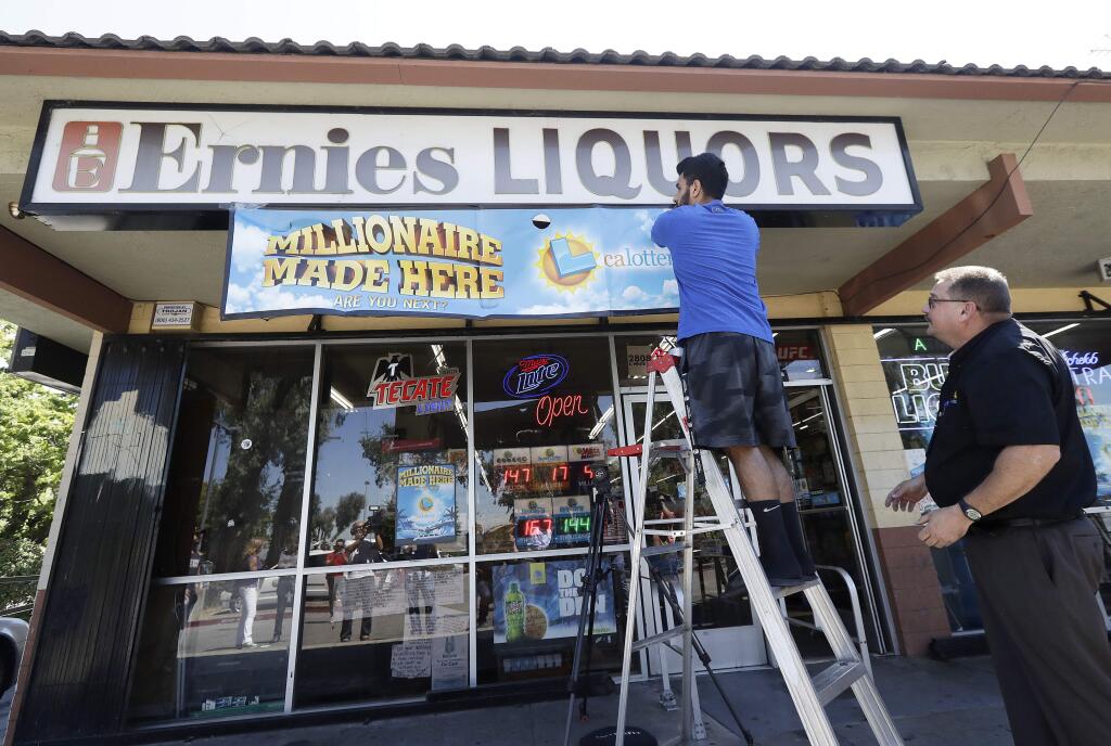 File - In this July 25, 2018 file photo, California Lottery official Mike Neis, right, watches as Amol Sachdev hangs a sign over his family's store Ernie's Liquors where a Mega Millions lottery ticket worth more than $500 million was sold in San Jose, Calif. The California Lottery says last month's $543 million Mega Millions jackpot was won by 11 members of an office pool that played on a whim. A Lottery statement Friday, Aug. 3, 2018, says Roland Reyes and the others showed up at its Hayward office on the east side of San Francisco Bay the day after the July 24 draw. The members declined to say where they work, other than that it is in the financial industry and plan to keep their jobs. They declined to speak to media or be photographed. (AP Photo/Jeff Chiu, File)