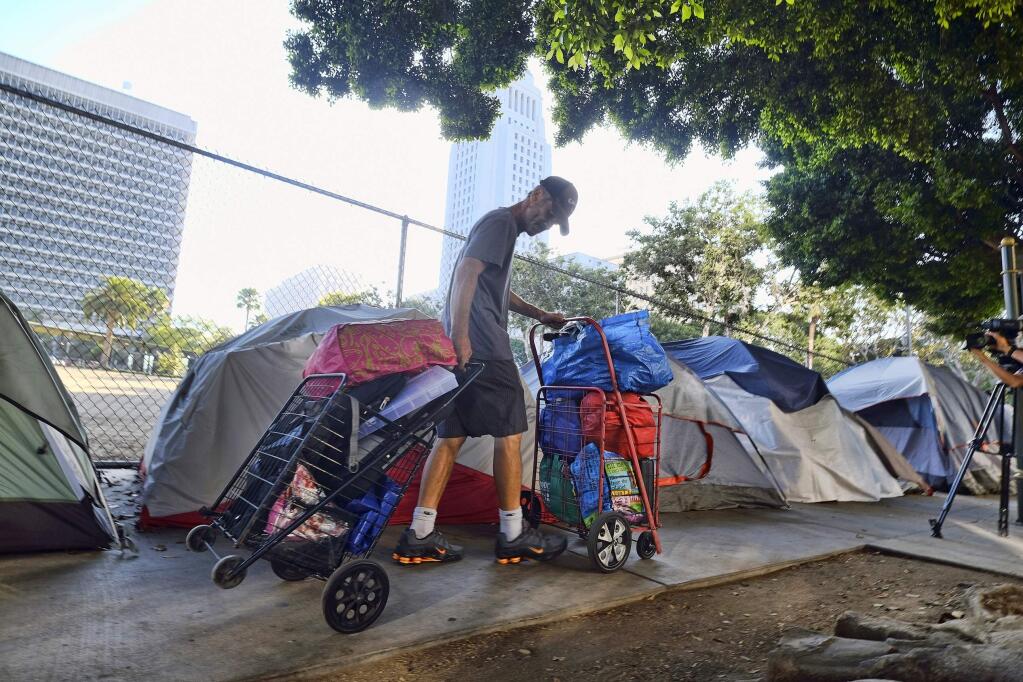 FILE - In this July 1, 2019 file photo, a homeless man moves his belongings from a street near Los Angeles City Hall, background, as crews prepared to clean the area. A proposal to restrict where homeless people may camp around Los Angeles drew protest at a City Council meeting from demonstrators who fear the rules would criminalize homelessness. Council members began discussion Tuesday, Sept. 24, 2019, on proposed changes to the city's code that would prevent people from sleeping near sensitive areas such as schools, or blocking right-of-ways like driveways and loading docks. (AP Photo/Richard Vogel, File)