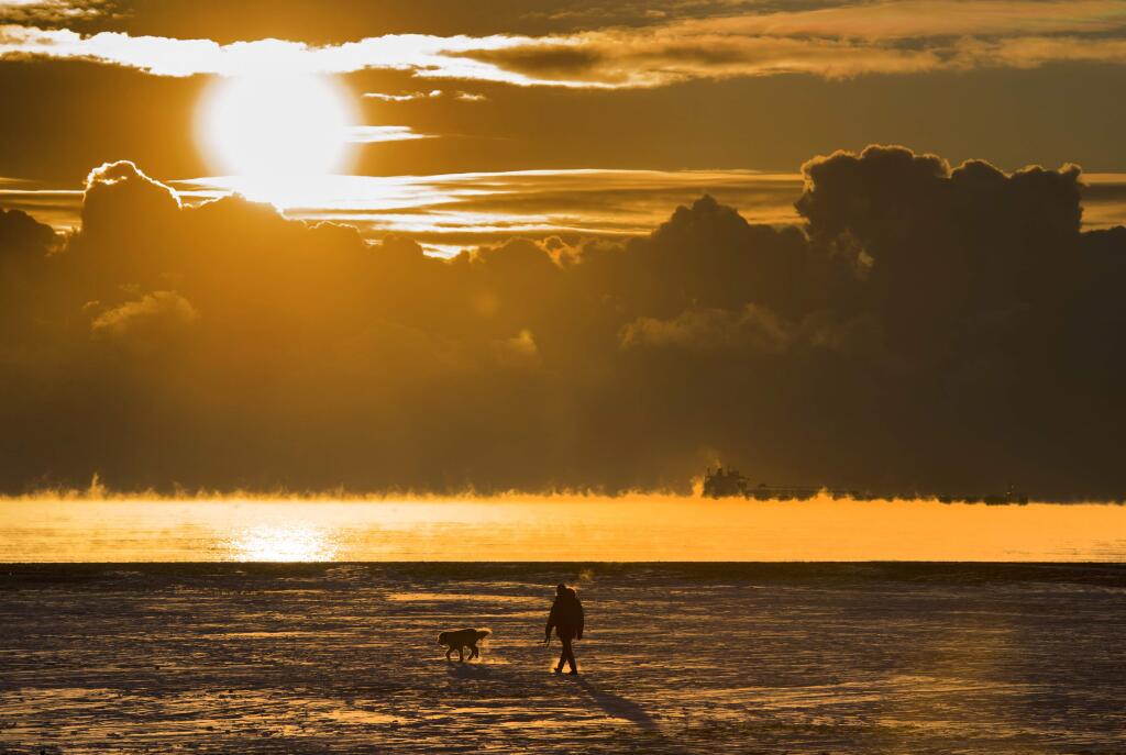 FILE - In this Wednesday, Dec. 27, 2017 file photo, a man walks his dog across the snow-covered beach while a cargo ship sits in the steaming fog of Lake Ontario in Toronto. According to a report released on Thursday, Jan. 18, 2018, U.S. and British scientists calculate that 2017 wasn't the hottest year on record, but close and unusually warm for no El Nino cooking the books. (Frank Gunn/The Canadian Press via AP)