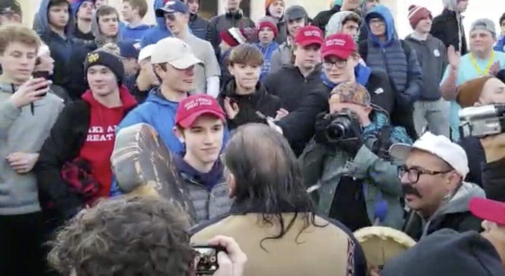 In this Friday, Jan. 18, 2019 image made from video provided by the Survival Media Agency, a teenager wearing a 'Make America Great Again' hat, center left, stands in front of an elderly Native American singing and playing a drum in Washington. The Roman Catholic Diocese of Covington in Kentucky is looking into this and other videos that show youths, possibly from the diocese's all-male Covington Catholic High School, mocking Native Americans at a rally in Washington. (Survival Media Agency via AP)
