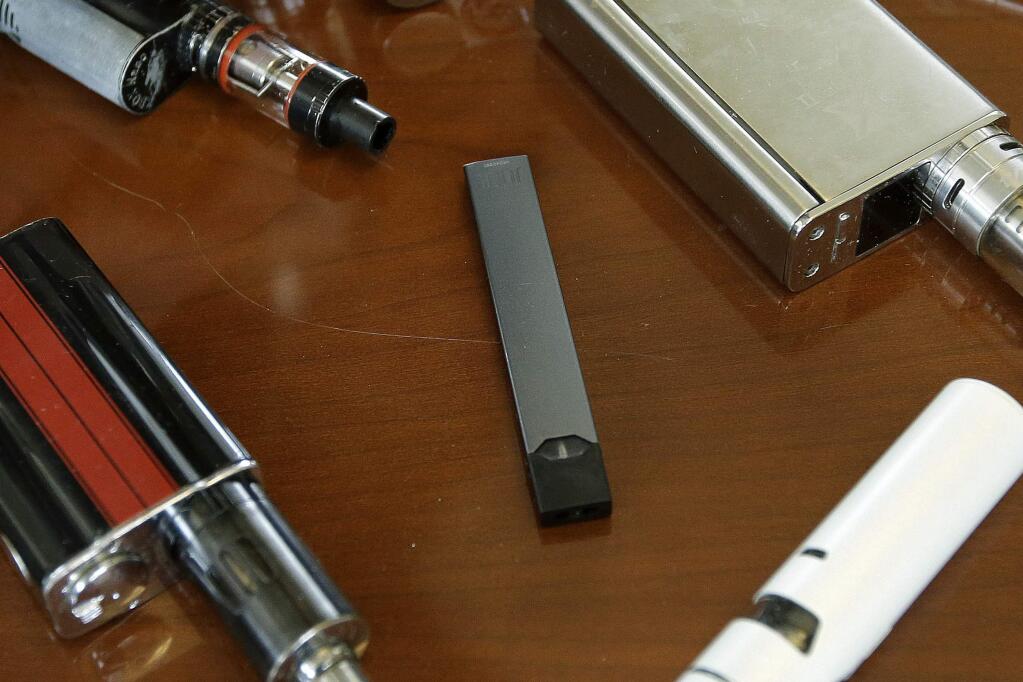 FILE - This Tuesday, April 10, 2018 file photo shows vaping devices, including a Juul, center, that were confiscated from students at a high school in Marshfield, Mass. On Tuesday, Nov. 13, 2018, San Francisco-based Juul Labs Inc. announced it had stopped filling orders for its mango, fruit, creme and cucumber pods but not menthol and mint. It will sell all flavors through its website and limit sales to those 21 and older. (AP Photo/Steven Senne)