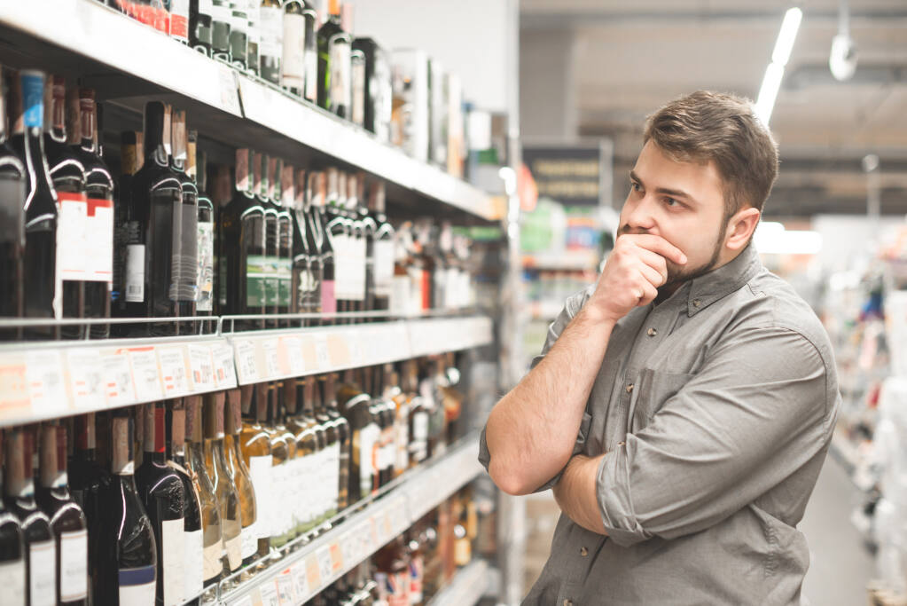 Bearded white young consumer holds his chin while intently looking at shelves of wine bottles in a supermarket.