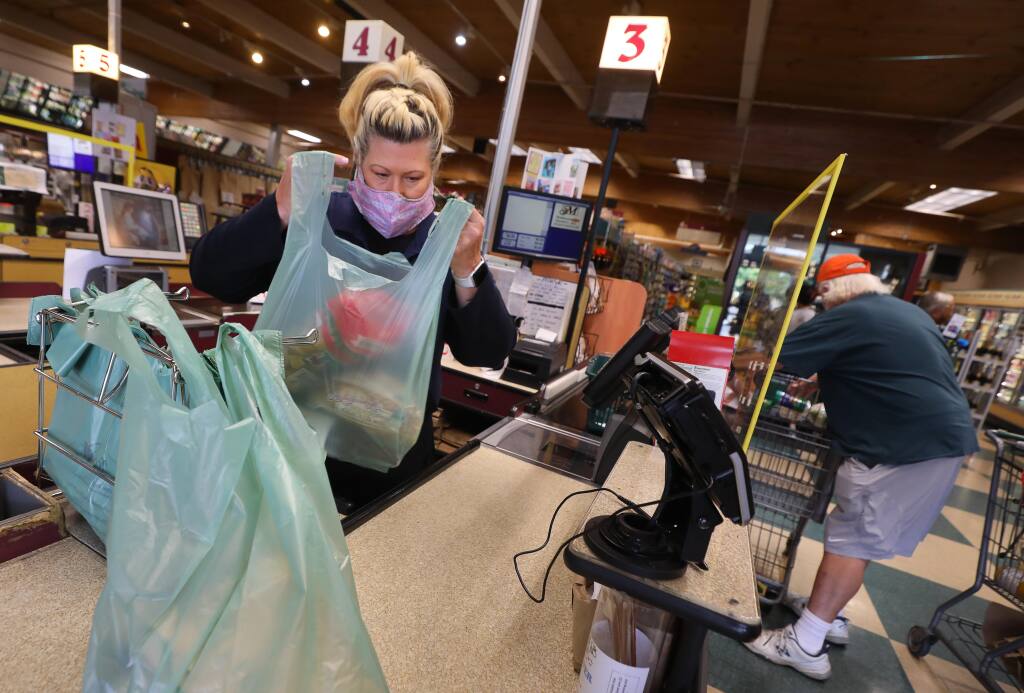 Cashier Michelle Paull bags a customer's purchases into plastic bags at Molsberry's Market in Santa Rosa on Thursday, July 16, 2020.  Grocery stores are beginning to allow the use reusable bags again, but customers are required to bag their own purchases, and refrain from placing personal bags onto high-touch surfaces such as checkout counters and conveyor belts.(Christopher Chung/ The Press Democrat)