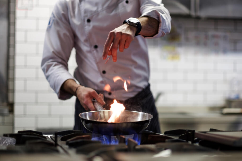 A kitchen cook in a white culinary uniform and black apron drops ingredients into a pan over a blue natural gas flame on the stove, while a yellow flame rise from inside the pan.