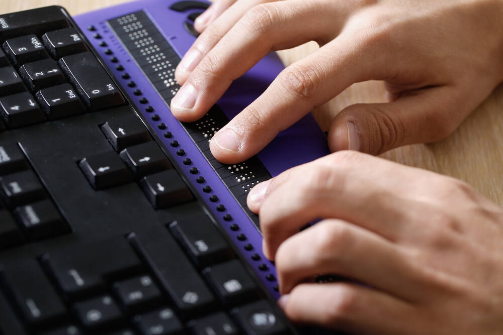 A blind person uses a computer keyboard with a braille display of screen content.