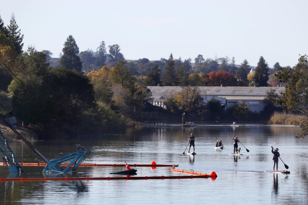 Paddle boarders and kayakers row past containment booms surrounding a barge and crane that is partially submerged and blocking 3/4 of the Petaluma River near Heritage Salvage in November. (BETH SCHLANKER/ The Press Democrat)