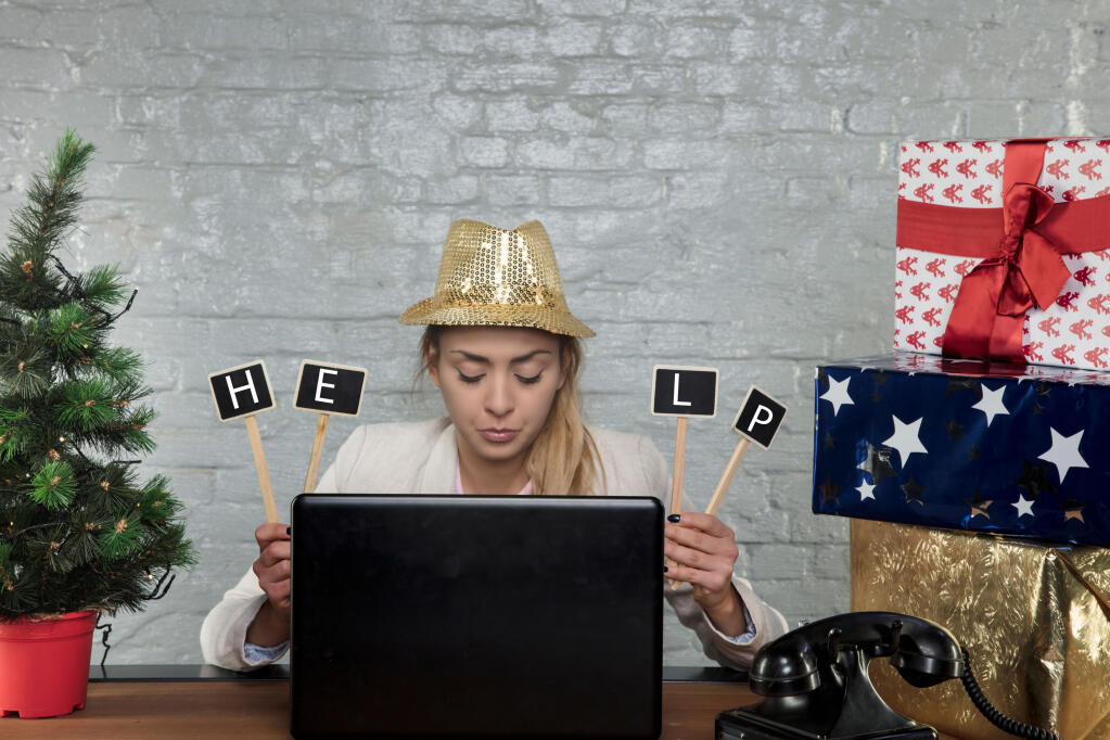 An unhappy woman wearing a sheer gold-sequin hat with a brim sits at an office desk surrounded by a tabletop Christmas tree and wrapped gift packages while looking down at her laptop keyboard and holding up four dowels with attached white letters on black backgrounds spelling "help." The employee is working instead of celebrating at the company party.