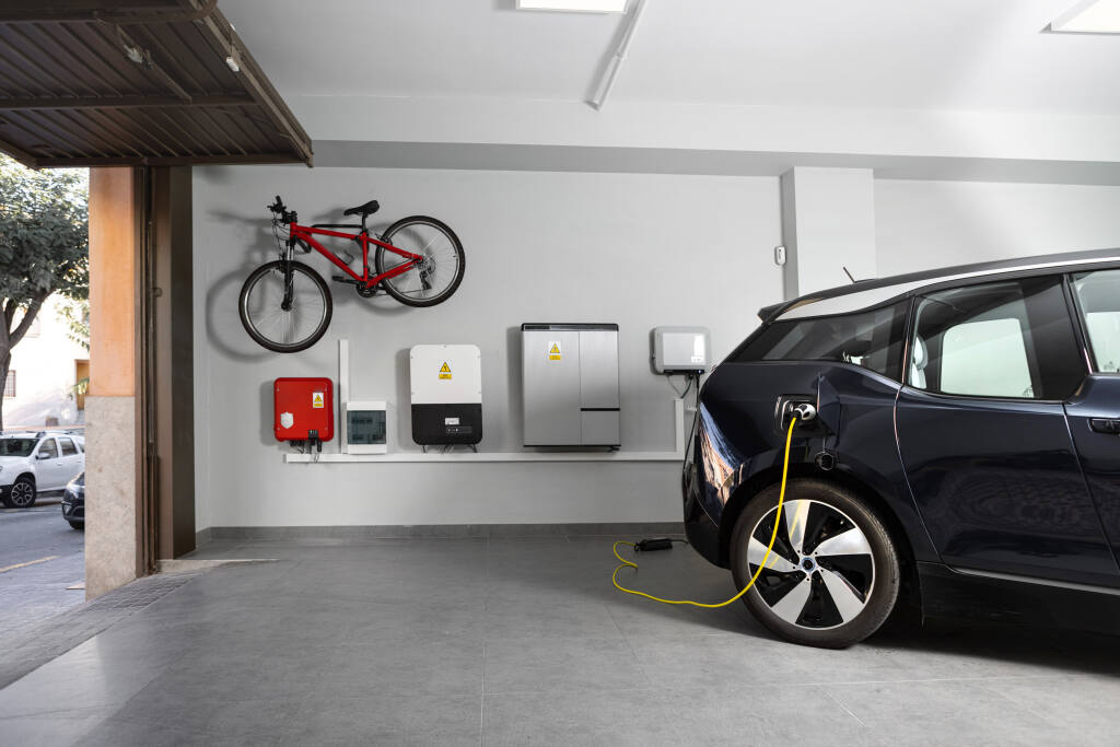 An electric vehicle is plugged into a charging unit inside a home garage. The garage door is shown open, and a bicycle is hanging above some of the wall-mounted charging equipment.