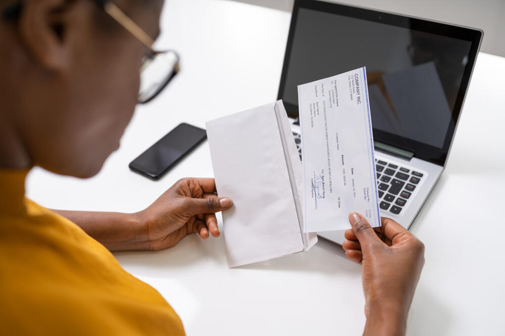 A Black businesswoman looks at a paycheck pulled out of an envelope in front of a laptop and cellphone.