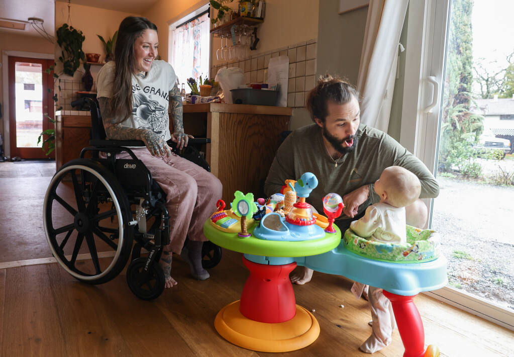 Jessie Marioni, left, watches her husband, Dan, play with their 8-month-old daughter, Maria, at their home in Sonoma on Friday, January 13, 2023.  On December 27, 2022, Jessie Marioni was carrying Maria on Black Sands Beach in Humboldt County when she was hit by a redwood log, that was pushed by a sleeper wave, that broke her pelvis. Dan Marioni came to their aid, with Maria coming out unscathed. (Christopher Chung/The Press Democrat)