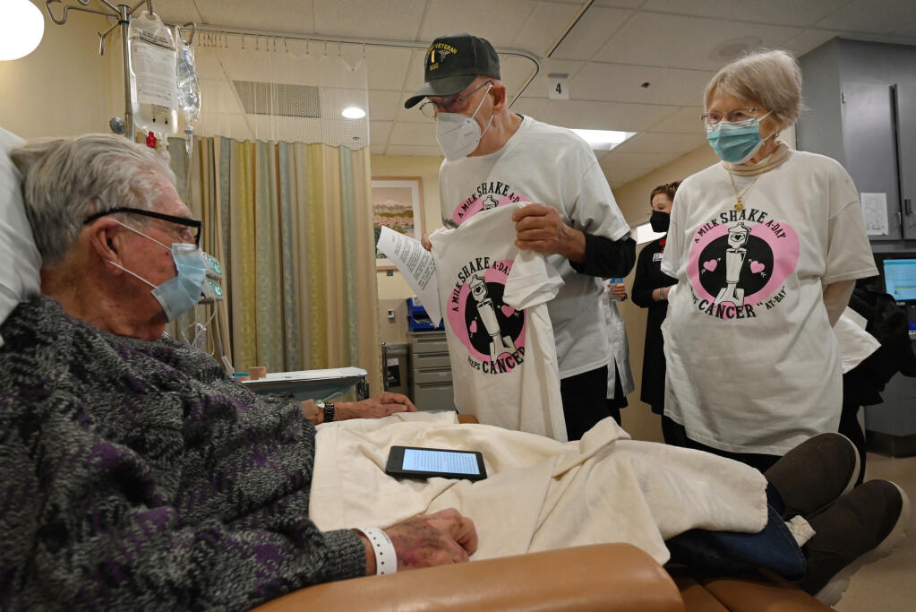 Rich Hoffman, center, and his wife Toby Hoffman, both 89, offer a T-shirt they made with a “Shake Cancer” milkshake recipe printed on the back to Michael Desky, 85, left, while he receives treatment at the Kaiser Permanente Oncology department in Santa Rosa on Tuesday, Jan. 31, 2023. Mrs. Hoffman just went through four years of cancer treatment at this facility and the couple wanted to offer up their support to fellow patients. (Erik Castro/For The Press Democrat)